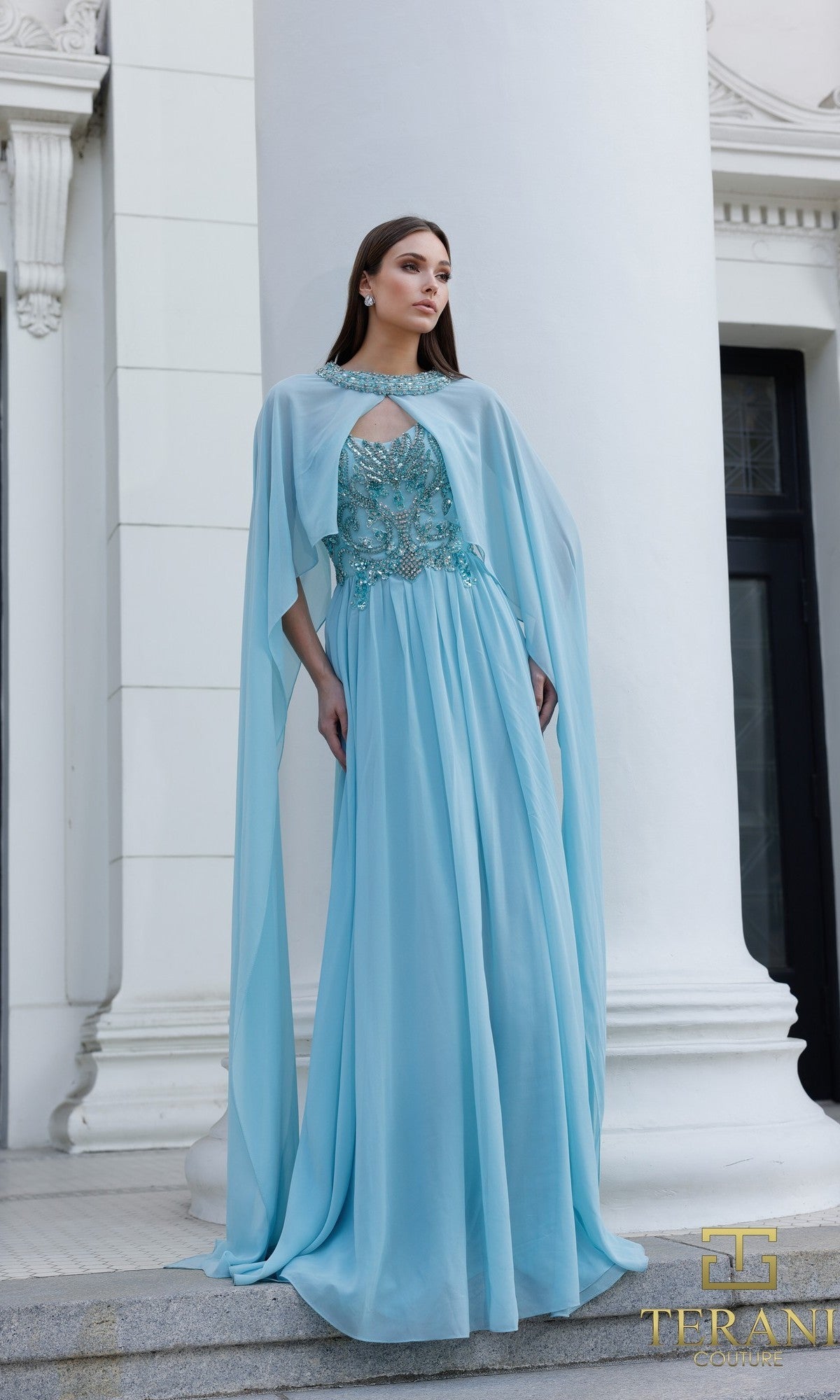 A woman stands before white columns wearing the Terani 261M6402 long formal dress in Sea Mist, featuring a strapless chiffon gown with a sheer cape and floral embroidered bodice.